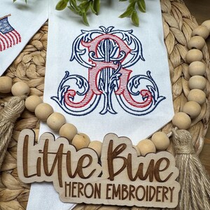 May include: A white fabric banner featuring an embroidered red and blue monogram letter R with ornate scrollwork. A wooden sign with the laser-cut text Little Blue HERON EMBROIDERY is placed over the banner, accompanied by a wooden bead garland.