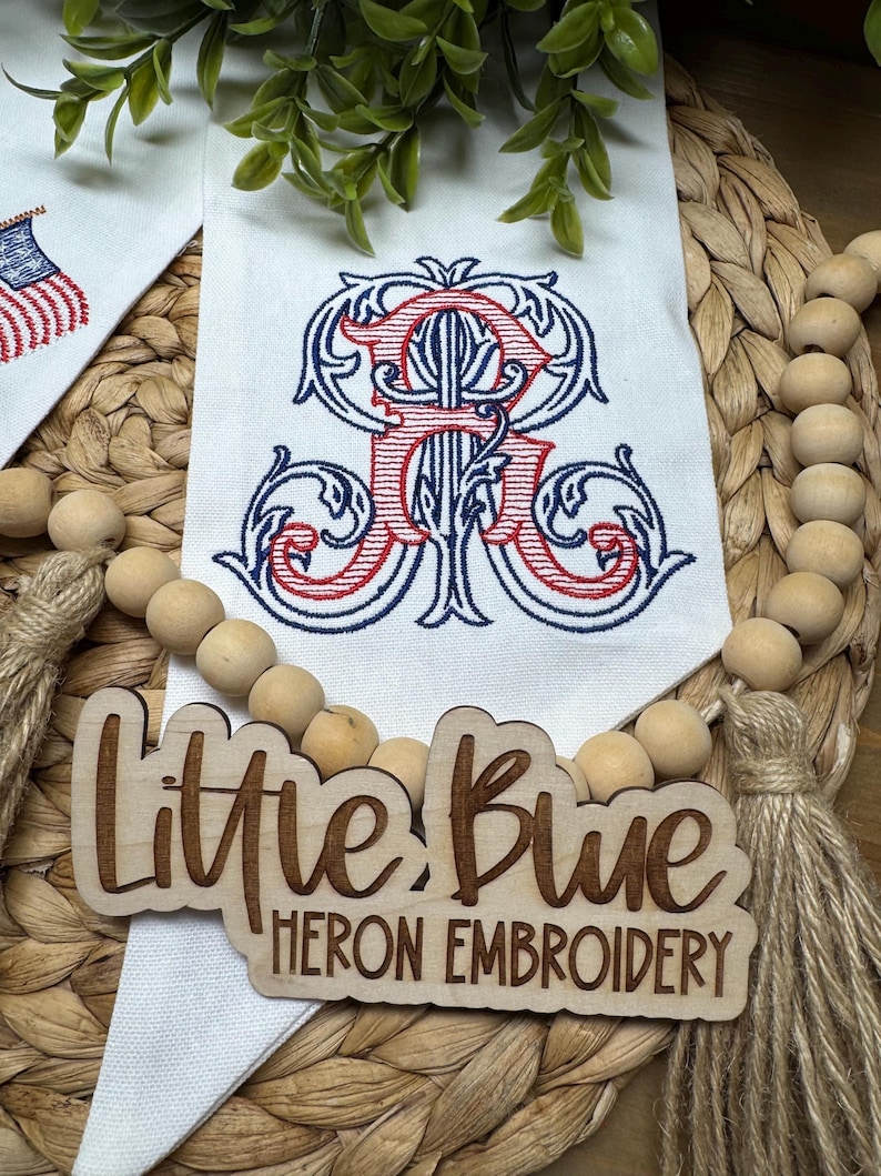 May include: A white fabric banner featuring an embroidered red and blue monogram letter R with ornate scrollwork. A wooden sign with the laser-cut text Little Blue HERON EMBROIDERY is placed over the banner, accompanied by a wooden bead garland.