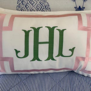 May include: White rectangular pillow with a pink geometric border and green embroidered initials JHL. The pillow is on a blue and white striped bedspread, with a patterned pillow in the background.