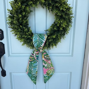 May include: A green wreath with a tropical print bow. The wreath is made of artificial greenery and the bow is made of fabric with a pink, green, and blue pattern.