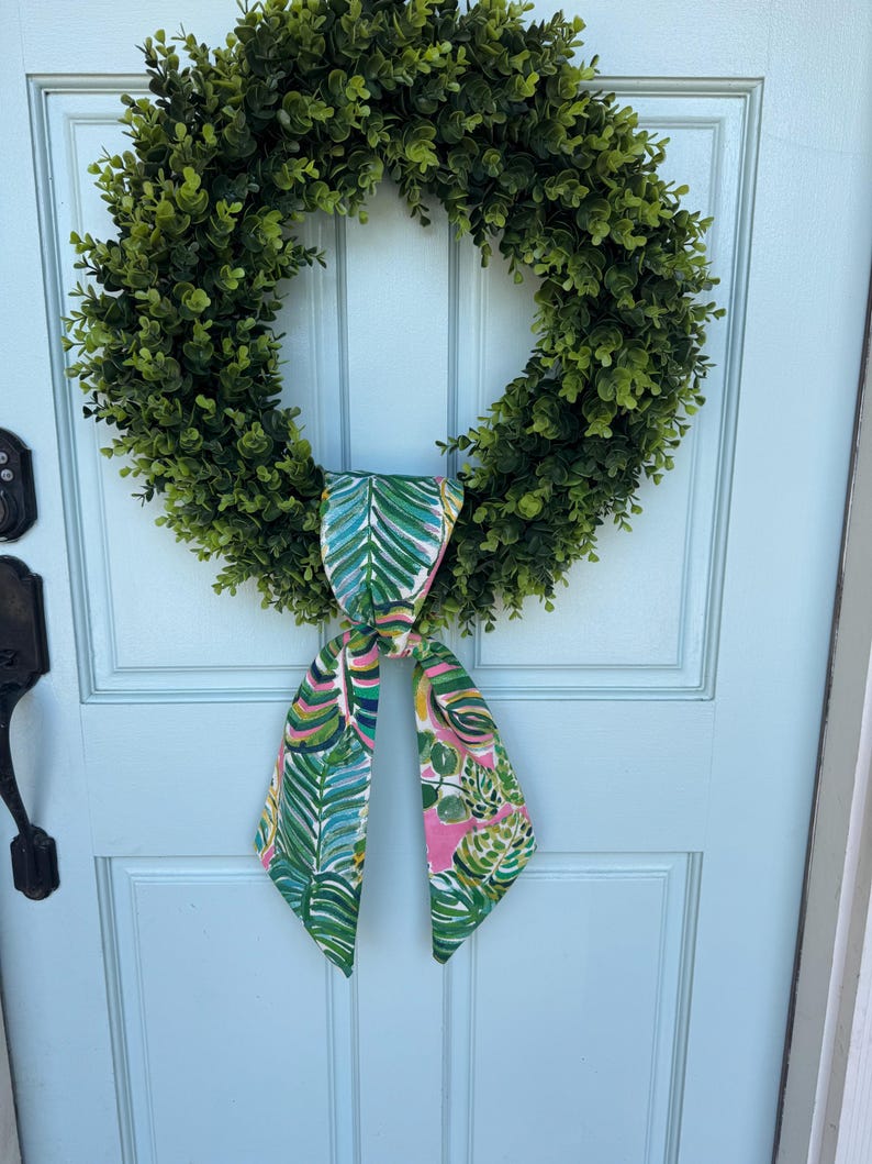 May include: A green wreath with a tropical print bow. The wreath is made of artificial greenery and the bow is made of fabric with a pink, green, and blue pattern.