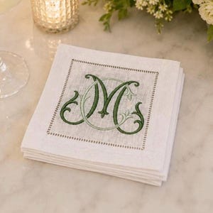 Può includere: Una pila di tovaglioli quadrati in lino bianco con un monogramma ricamato della lettera M al centro in filo verde scuro. Il monogramma è circondato da un bordo decorativo di piccoli punti verdi, creando un design pulito e strutturato.