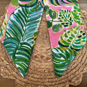 May include: A pink fabric with a green and white tropical leaf pattern. The fabric is folded in half and the edges are frayed.