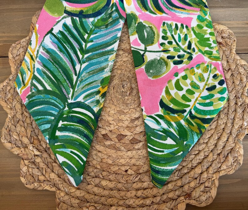 May include: A pink fabric with a green and white tropical leaf pattern. The fabric is folded in half and the edges are frayed.
