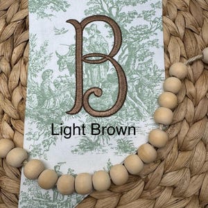 May include: A white fabric pennant featuring a green toile pattern of figures and trees, centered with a large embroidered letter B in dark brown thread. The text Light Brown is printed below the letter. A string of natural wood beads is attached.