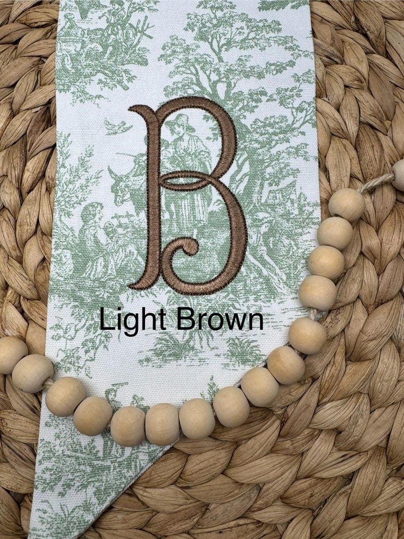 May include: A white fabric pennant featuring a green toile pattern of figures and trees, centered with a large embroidered letter B in dark brown thread. The text Light Brown is printed below the letter. A string of natural wood beads is attached.