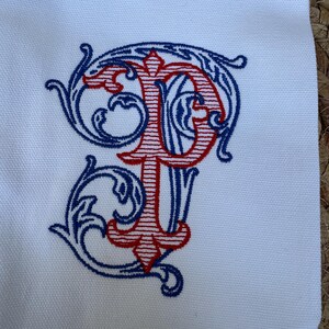 May include: A white fabric surface featuring an embroidered monogram of the letters P and T. The letter P is stitched in red with horizontal lines, while the letter T is intertwined with the P in blue, surrounded by decorative blue scrollwork.