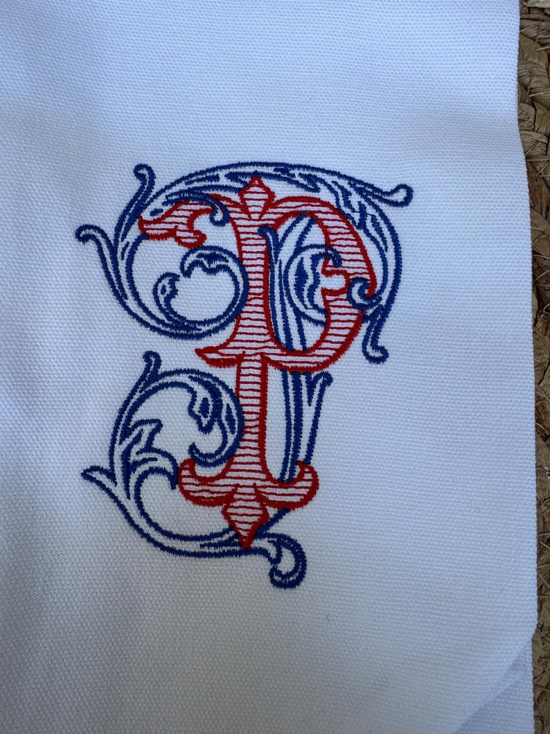 May include: A white fabric surface featuring an embroidered monogram of the letters P and T. The letter P is stitched in red with horizontal lines, while the letter T is intertwined with the P in blue, surrounded by decorative blue scrollwork.