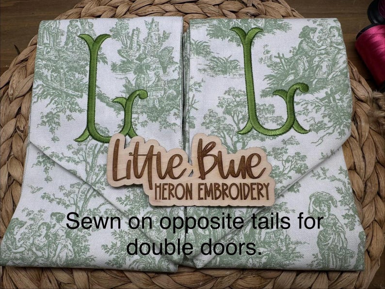 May include: Two white fabric panels featuring a green toile pattern and large embroidered green letters. A wooden sign reads Little Blue Heron Embroidery. Text on the image states: Sewn on opposite tails for double doors.
