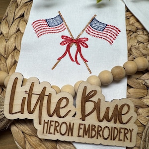 May include: A white fabric banner featuring an embroidered design of two crossed American flags tied with a red bow. A wooden sign with the laser-engraved text "Little Blue HERON EMBROIDERY" is placed over the banner, accompanied by a string of wooden beads.