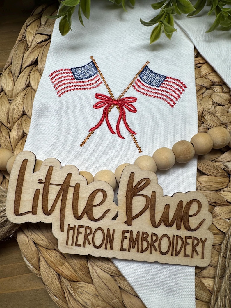 May include: A white fabric banner featuring an embroidered design of two crossed American flags tied with a red bow. A wooden sign with the laser-engraved text "Little Blue HERON EMBROIDERY" is placed over the banner, accompanied by a string of wooden beads.