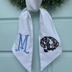 May include: White fabric bow with blue embroidered letter M on one side and a black embroidered dog silhouette on the other side.