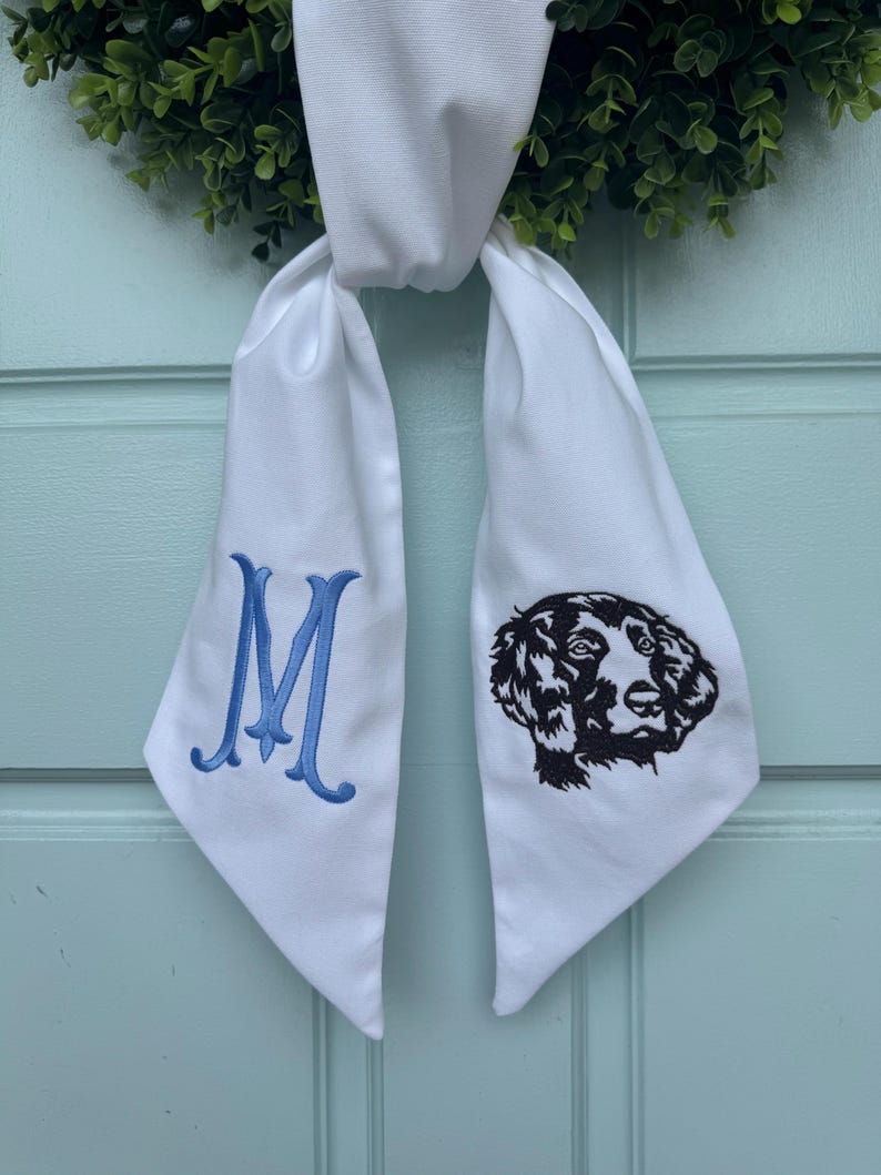 May include: White fabric bow with blue embroidered letter M on one side and a black embroidered dog silhouette on the other side.