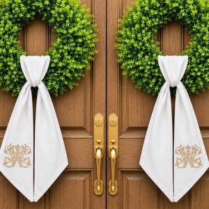 May include: Two green wreaths hang on a brown wooden door, each adorned with a white fabric accent. The fabric is embroidered with a gold monogram. Gold door handles and hardware complete the look, creating a welcoming entrance.