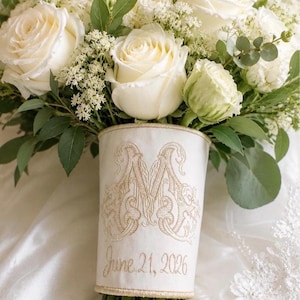 K&ouml;nnte beinhalten: Ein Brautstrau&szlig; aus wei&szlig;en Rosen und gr&uuml;nem Blattwerk, umwickelt mit einer cremefarbenen Stoffmanschette. Die Manschette ist mit einem goldenen Monogramm-Buchstaben M und dem Datum 21. Juni 2026 in Schreibschrift mit Rankenmotiven bestickt.