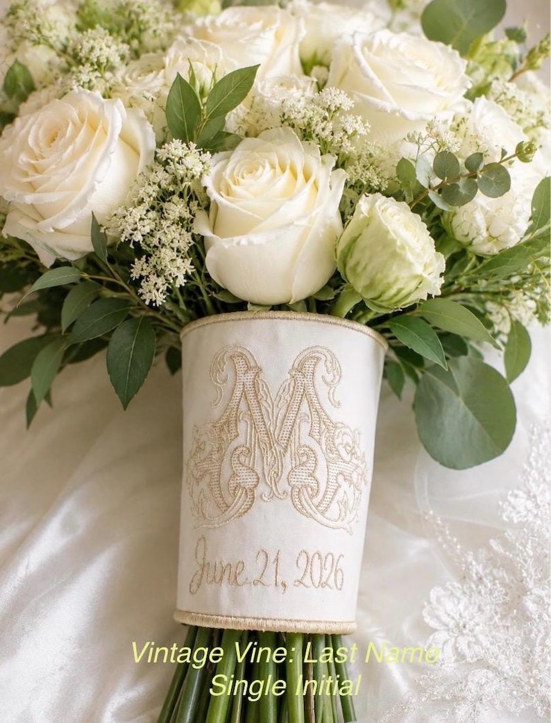 K&ouml;nnte beinhalten: Ein Brautstrau&szlig; aus wei&szlig;en Rosen und gr&uuml;nem Blattwerk, umwickelt mit einer cremefarbenen Stoffmanschette. Die Manschette ist mit einem goldenen Monogramm-Buchstaben M und dem Datum 21. Juni 2026 in Schreibschrift mit Rankenmotiven bestickt.