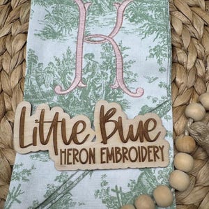 May include: A folded fabric napkin featuring a green and white toile pattern with a large embroidered letter K in flesh pink thread. A wooden sign with the laser-cut text Little Blue HERON EMBROIDERY is placed on top of the napkin.
