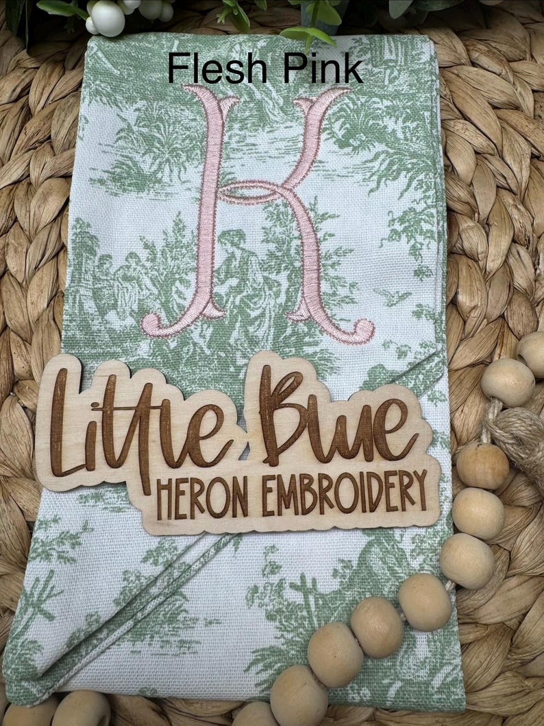 May include: A folded fabric napkin featuring a green and white toile pattern with a large embroidered letter K in flesh pink thread. A wooden sign with the laser-cut text Little Blue HERON EMBROIDERY is placed on top of the napkin.
