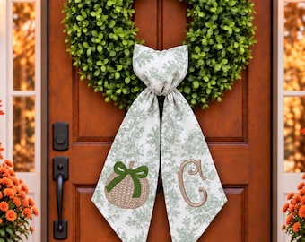 May include: A circular green boxwood wreath adorned with a long, hanging fabric ribbon. The ribbon features a light green botanical print, an embroidered tan pumpkin with a green bow, and a large embroidered tan letter C monogram.