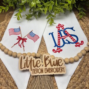 May include: Two white fabric pennants featuring machine embroidery. The left pennant displays two crossed American flags tied with a red bow. The right pennant features the letters US in red and blue. A wooden sign reads Little Blue Heron Embroidery.