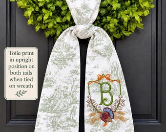 May include: A white fabric sash with a green toile print featuring a knotted centre and two hanging tails. One tail displays an embroidered green letter B inside a floral wreath with an orange bow and a colourful turkey at the base.