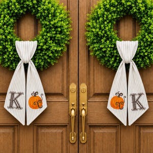 May include: Two circular green boxwood wreaths hanging on double wooden doors. Each wreath features a beige and white striped fabric ribbon tied at the bottom. The ribbons display a printed dark brown letter K and an orange pumpkin illustration.