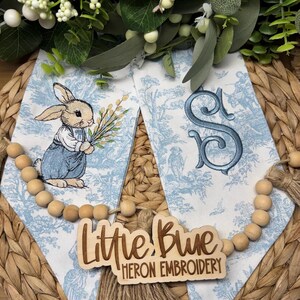 May include: Embroidered blue and white fabric with a bunny holding flowers and a monogram. The fabric is decorated with a toile pattern. A wooden sign reads "Little Blue Heron Embroidery". A wooden bead garland is draped across the fabric.
