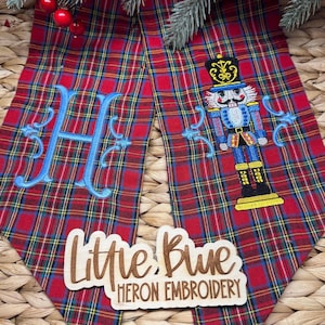 Könnte beinhalten: Roter karierter Weihnachtsbaumrock mit einem blau gestickten Buchstaben "H" und einem Nussknacker-Design. Der Rock hat ein Holzschild mit der Aufschrift "Little Blue Heron Embroidery".