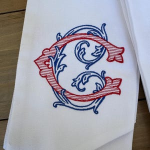 May include: A white fabric towel featuring an embroidered monogram of the letter G. The letter is designed with a red striped pattern and is surrounded by intricate blue scrollwork embroidery. The fabric has a visible woven texture.