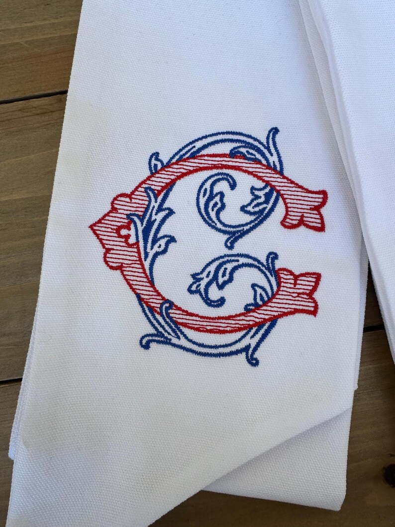 May include: A white fabric towel featuring an embroidered monogram of the letter G. The letter is designed with a red striped pattern and is surrounded by intricate blue scrollwork embroidery. The fabric has a visible woven texture.