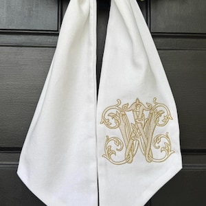 May include: White fabric door hanger with a gold embroidered monogram letter "W". The fabric is folded and tied at the top, with the monogram centered on the lower portion. The background is a black door.