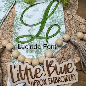 May include: A white fabric banner with a green toile pattern and a large embroidered green letter L. The text Lucinda Font is printed above a wooden cutout sign that reads Little Blue HERON EMBROIDERY, accented by a string of natural wooden beads.