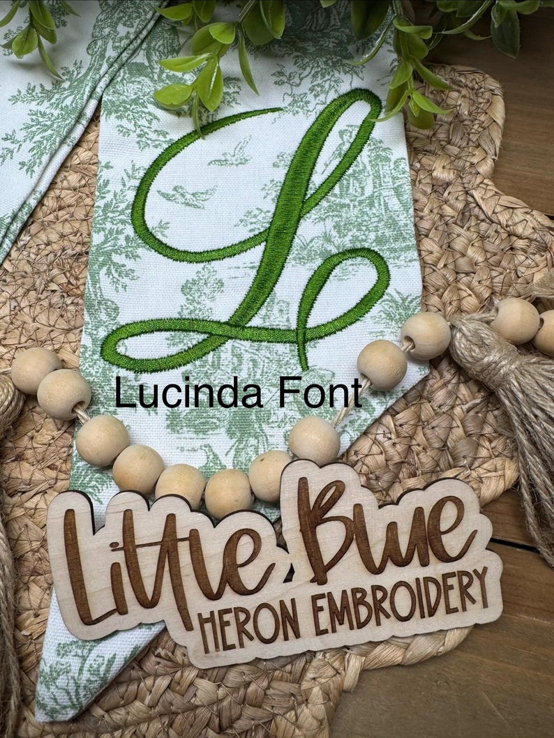 May include: A white fabric banner with a green toile pattern and a large embroidered green letter L. The text Lucinda Font is printed above a wooden cutout sign that reads Little Blue HERON EMBROIDERY, accented by a string of natural wooden beads.