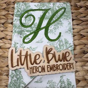 May include: A white fabric towel featuring a green toile pattern and a large embroidered green letter H. A light-colored wooden sign is placed over the towel with the laser-cut text Little Blue HERON EMBROIDERY in a script and block font.