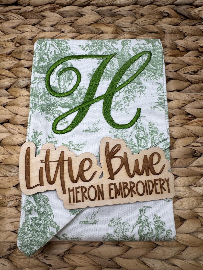 May include: A white fabric towel featuring a green toile pattern and a large embroidered green letter H. A light-colored wooden sign is placed over the towel with the laser-cut text Little Blue HERON EMBROIDERY in a script and block font.