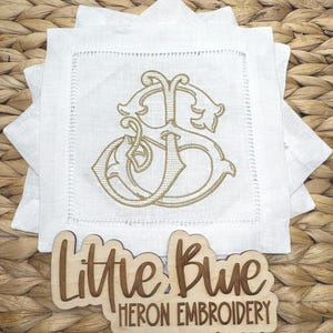May include: A stack of square white linen napkins featuring a gold embroidered monogram of the letters S and J in the center. A wooden sign in front displays the text Little Blue HERON EMBROIDERY in a script and block font combination.