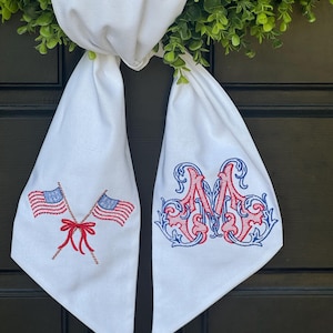 May include: A white fabric door hanger featuring two embroidered American flags crossed with a red bow on the left side and an ornate, decorative letter M embroidered in red and blue thread on the right side, hanging from a green wreath.