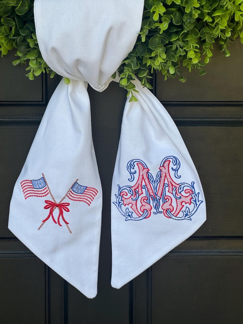 May include: A white fabric door hanger featuring two embroidered American flags crossed with a red bow on the left side and an ornate, decorative letter M embroidered in red and blue thread on the right side, hanging from a green wreath.