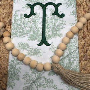 May include: A white fabric pouch featuring a green toile pattern of figures and trees, embroidered with a dark green letter T. The text Deep Green is printed above the letter. A string of natural wooden beads with a jute tassel is draped across.