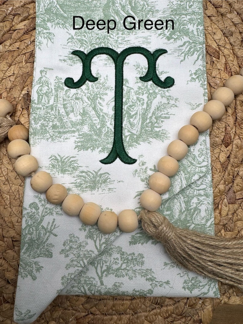 May include: A white fabric pouch featuring a green toile pattern of figures and trees, embroidered with a dark green letter T. The text Deep Green is printed above the letter. A string of natural wooden beads with a jute tassel is draped across.