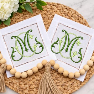 May include: Two square white linen cocktail napkins featuring a decorative green embroidered monogram letter M with intricate scrollwork details and a hemstitched border. The napkins are displayed with a natural wooden bead garland.