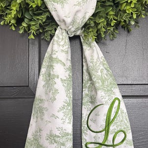 May include: A white fabric ribbon featuring a green toile de jouy pattern of pastoral scenes and a large, embroidered green script letter L. The ribbon is tied in a knot and hangs down with two pointed ends, layered over a green artificial wreath.