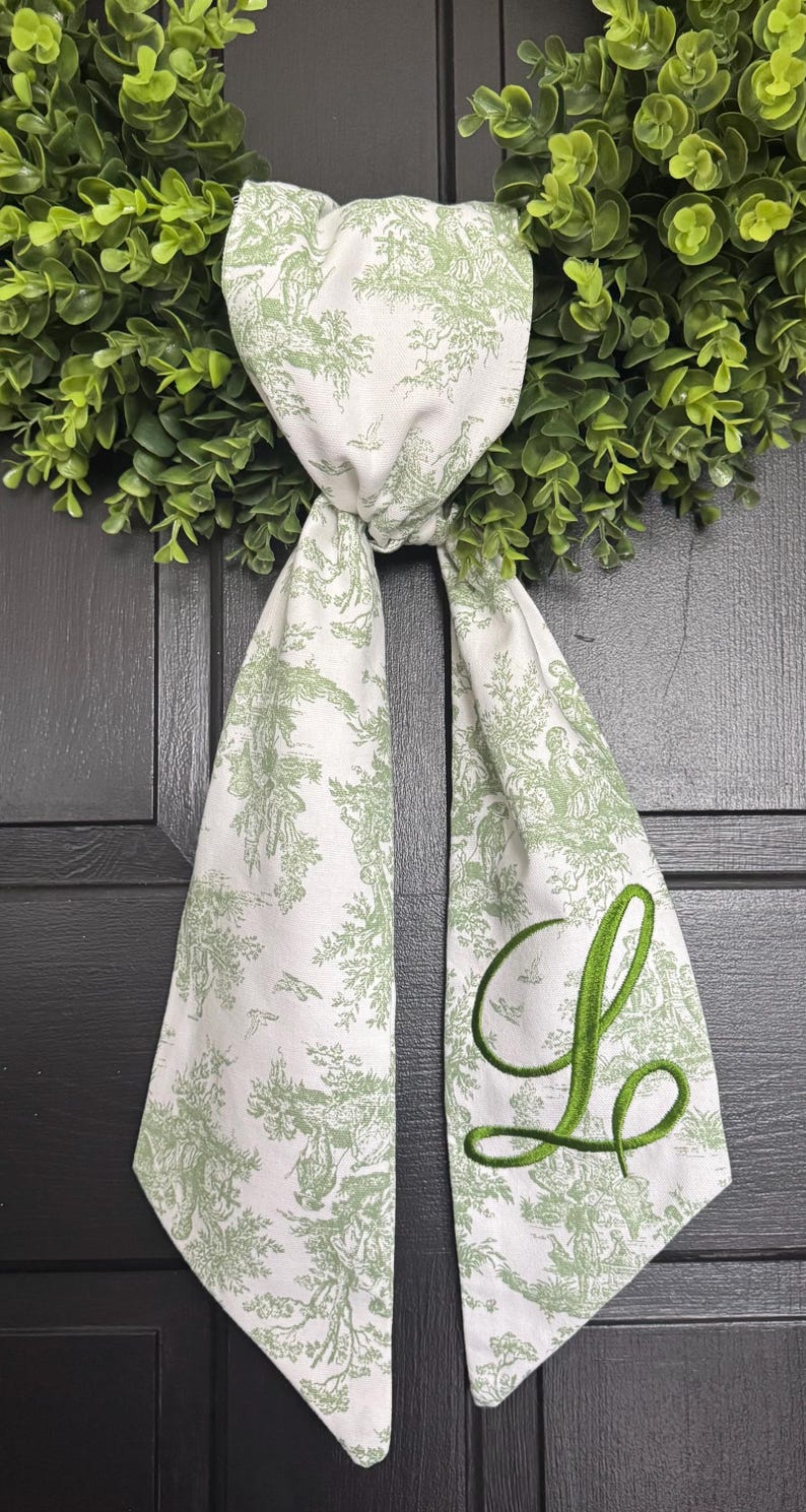 May include: A white fabric ribbon featuring a green toile de jouy pattern of pastoral scenes and a large, embroidered green script letter L. The ribbon is tied in a knot and hangs down with two pointed ends, layered over a green artificial wreath.