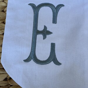 May include: A white fabric banner with a large, gray embroidered letter "E" in a decorative script font.