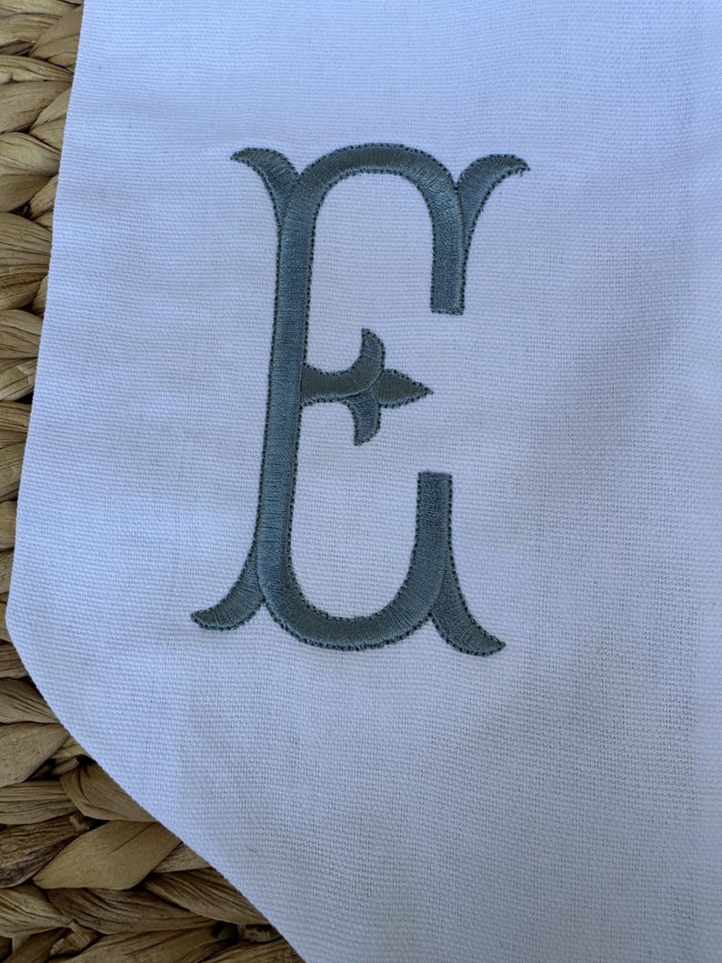 May include: A white fabric banner with a large, gray embroidered letter "E" in a decorative script font.