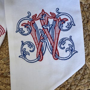 May include: A white fabric item featuring an embroidered monogram of the letter W. The design is rendered in red and blue thread with intricate, swirling filigree details surrounding the central letter, creating a decorative and ornate appearance.