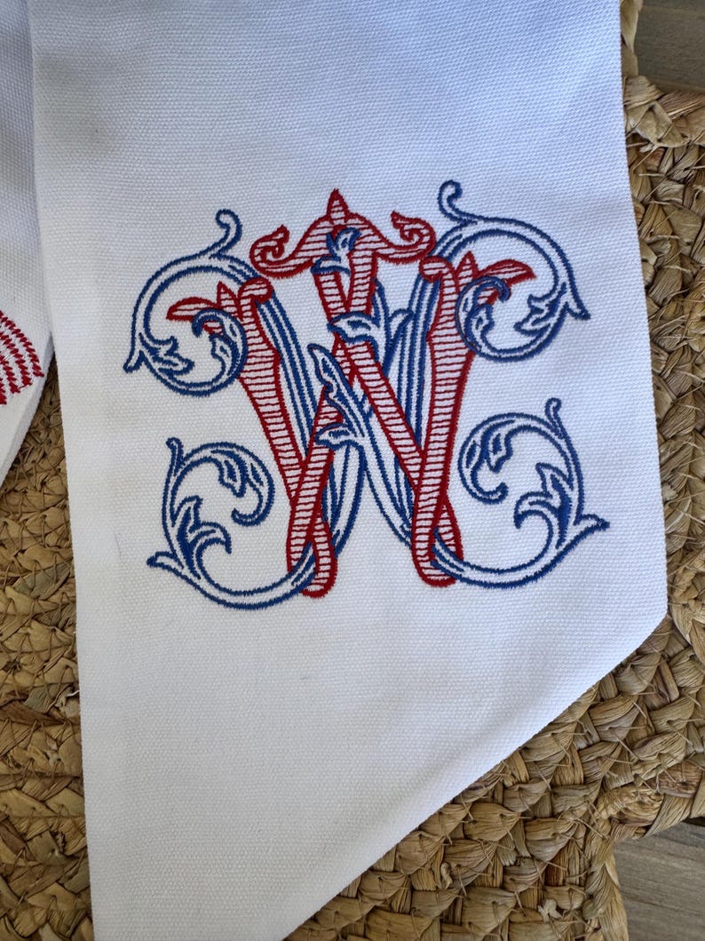 May include: A white fabric item featuring an embroidered monogram of the letter W. The design is rendered in red and blue thread with intricate, swirling filigree details surrounding the central letter, creating a decorative and ornate appearance.