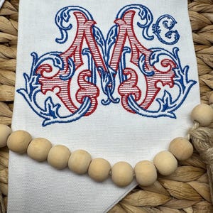 May include: A white fabric banner featuring an embroidered monogram of the letter M in red and blue thread with ornate scrollwork details. A string of natural light-colored wooden beads is draped horizontally across the lower section of the banner.