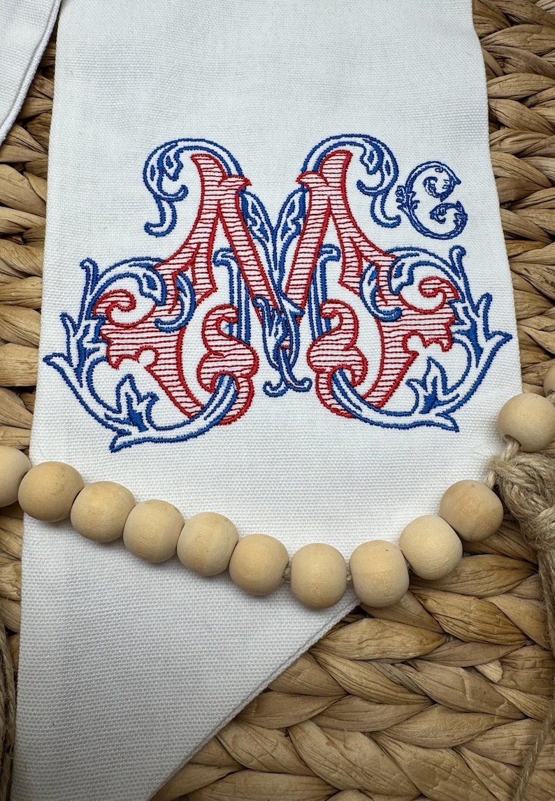 May include: A white fabric banner featuring an embroidered monogram of the letter M in red and blue thread with ornate scrollwork details. A string of natural light-colored wooden beads is draped horizontally across the lower section of the banner.