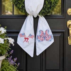 May include: A white fabric door hanger featuring two embroidered American flags crossed with a red bow on the left tail and a large, ornate, red and blue embroidered letter M on the right tail, draped over a green wreath on a black door.